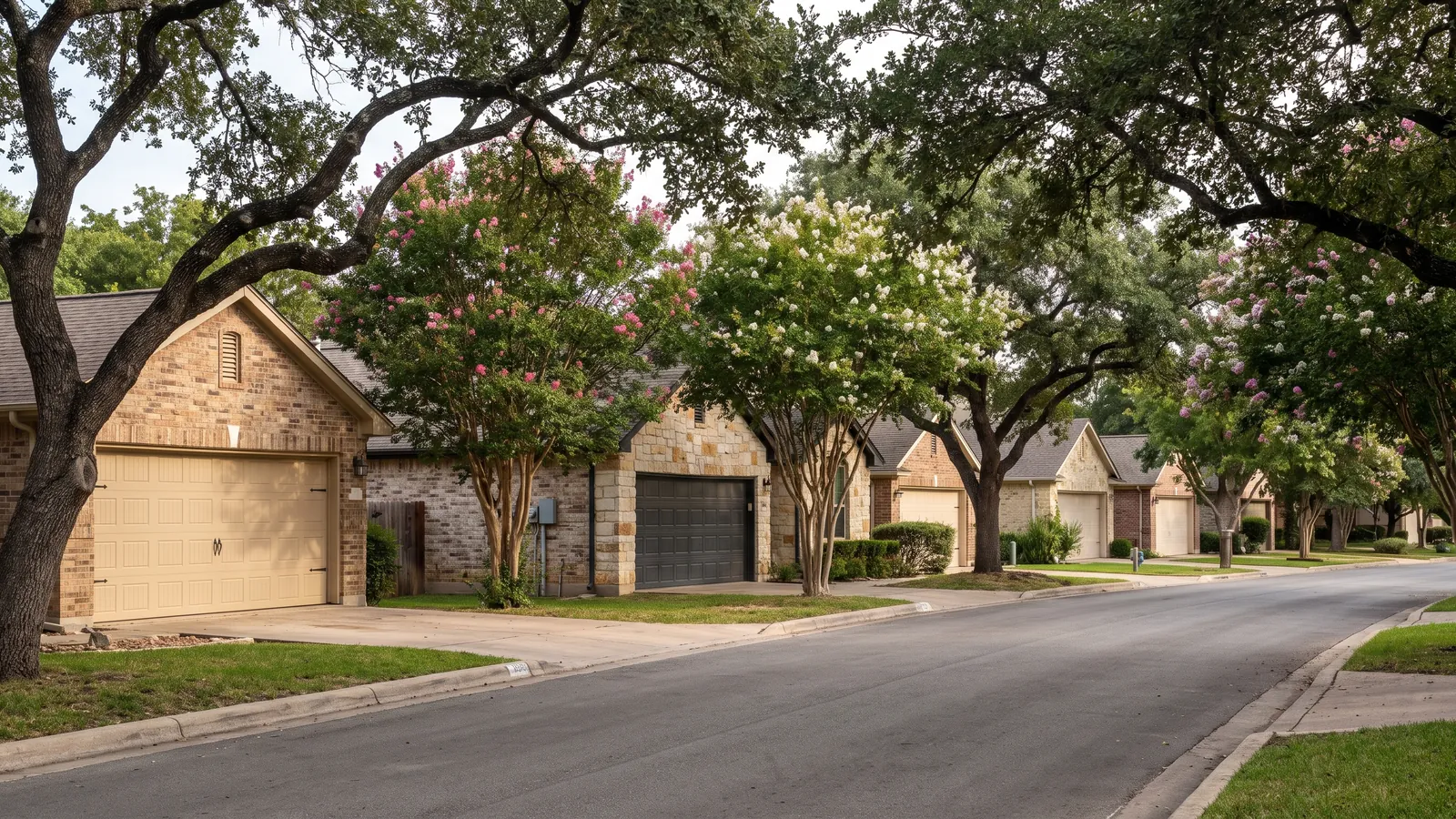 Quiet Austin neighbourhood street lined with one-story brick and limestone homes, each with a two-car garage door, live oaks and crepe myrtles along the curb